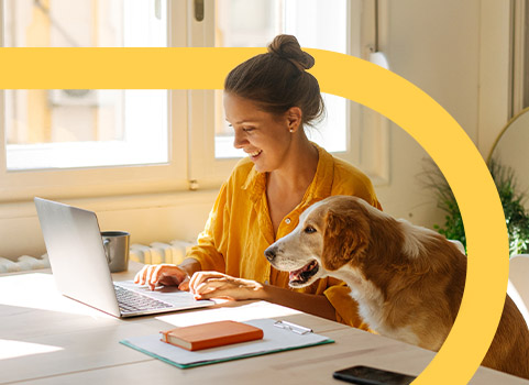 Woman in yellow shirt using laptop with dog beside her.