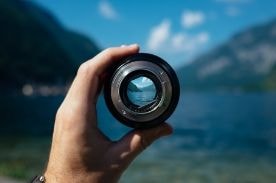 person holding camera lens near lake.