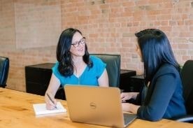 Two women facing one another with laptop on table in front of them.