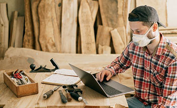 Man wearing mask working on laptop at wooden table with carpentry tools.