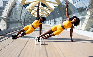 Two women wearing yellow doing raised arm side planks on a bridge outside.