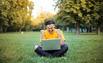 Man sitting in grass with an open laptop on his lap.