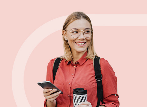 Young woman wearing glasses holding a smart phone in one hand and a travel cup of coffee in the other on a light pink background.