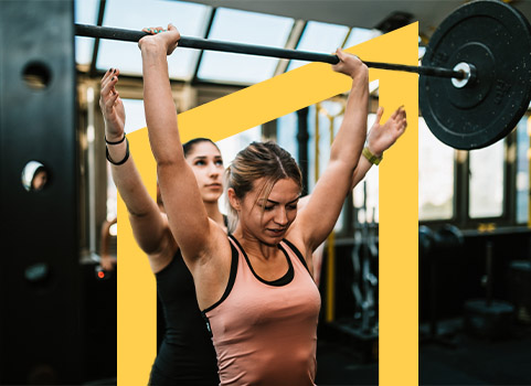 Woman in pink tank top lifting weight with woman in black spotting behind her.