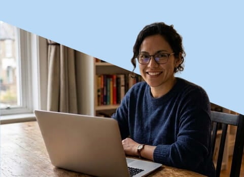 Adult women with dark hair and glasses sitting at kitchen table with silver laptop.