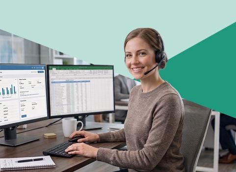 woman in brown sweater and headset sitting at desk reviewing reports on computer monitors.