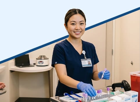 Phlebotomy technician wearing navy blue scrubs, a hospital name badge, and blue sterile gloves holding vials for collecting blood from patients in an exam room.