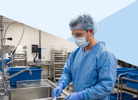 Man in scrubs, face mask, gloves, and hairnet sterilizing medical equipment.
