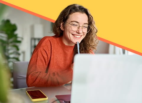 smiling woman in an orange sweater holding a pen and looking at a laptop.