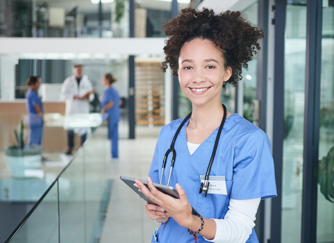 A woman with light skin and dark, curly hair wears scrubs and a stethoscope and holds a tablet