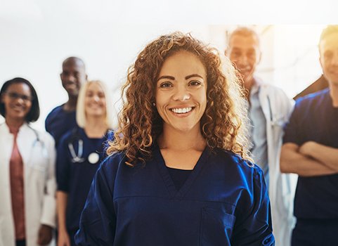 A woman with medium brown skin and curly brown hair wearing scrubs stands in the foreground; multiple people stand behind her wearing lab coats and scrubs and stethoscopes