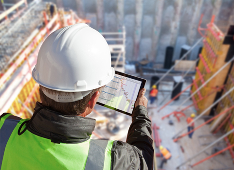 A person wearing a hardhat looks at a computer while overlooking a construction scene