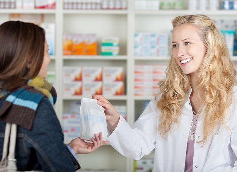 A woman with fair skin and blonde hair wears a lab coat and hands medication to a woman with medium skin and dark hair