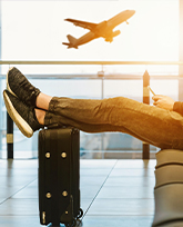 A person reclines in an airport lounge, watching a plane lift off through the airport window