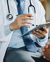 A person in a suit and tie wearing a lab coat and a stethoscope, holding an ipad and gesturing to another person with folded hands