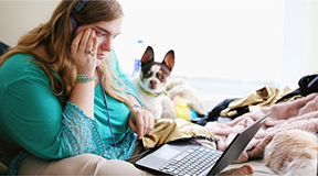 Girl studying on laptop with small dog in the background.