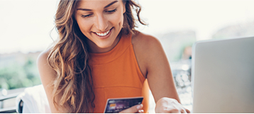 Girl in orange shirt looking at her phone while using a laptop outside.