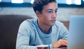 Teen boy in gray long sleeve shirt focusing on silver laptop.