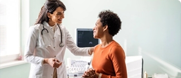 Healthcare worker in white lab coat with stethoscope speaking with a patient wearing an orange shirt in an exam room.