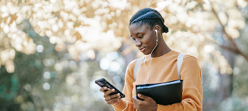 Teen girl outside in orange sweater with earbuds looking at smartphone.
