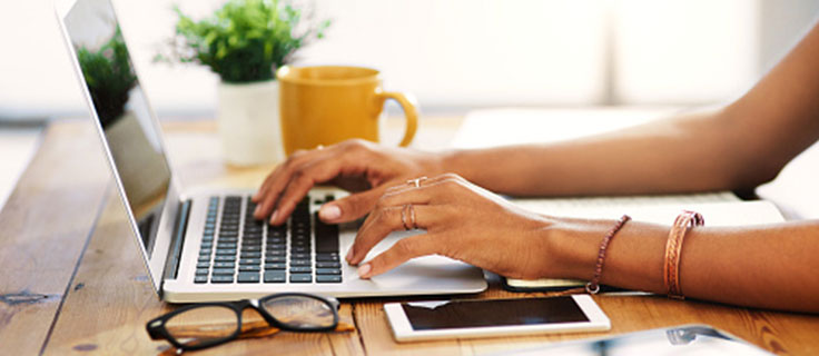 woman typing on a computer with coffee and a cell phone.