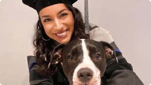 College student in graduation regalia holding black and white dog.