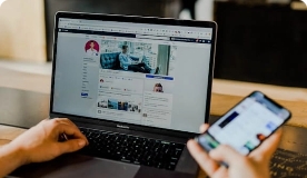Closeup of laptop showing Facebook page while a hand holds a smartphone.