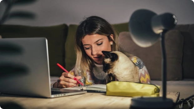 Girl taking notes with cat in front of laptop on coffee table in living room.