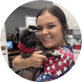 Penn Foster Vet Tech grad in red, white, and blue starred scrubs holding small dog in vet clinic. 