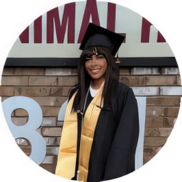 Penn Foster graduate in black regalia and yellow stole posing in front of business. 